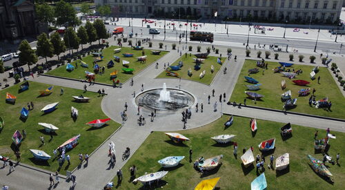 Großer Platz mit Brunnen in der Mitte. Ringsum auf dem Rasen große bunte Papierboote.